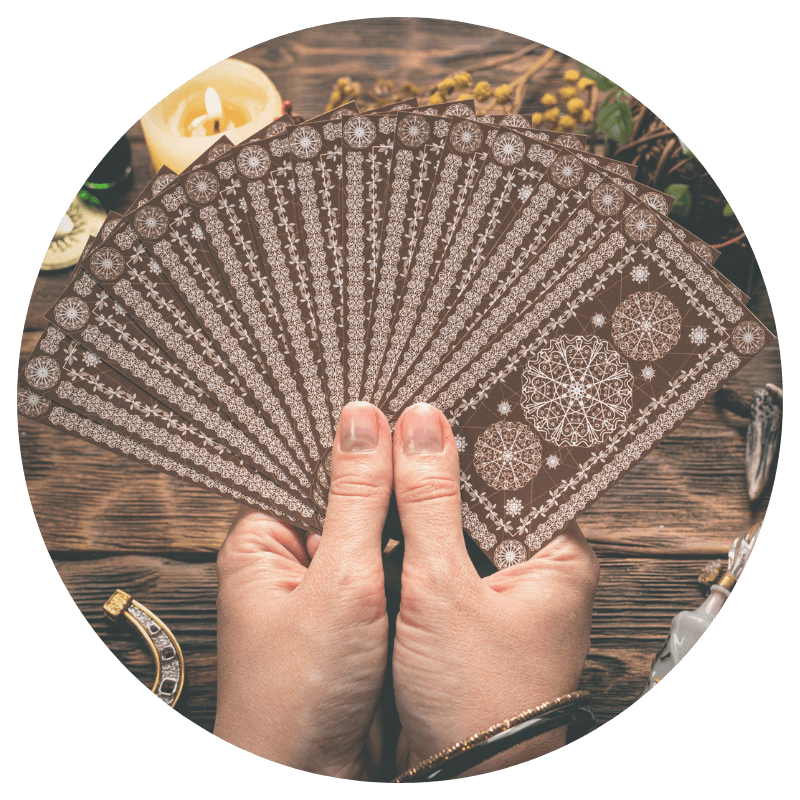 two white hands holding a fanned out deck of tarot cards over a wooden table